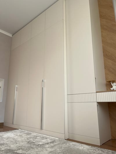The image shows a modern, built-in wardrobe in a room. The wardrobe has several tall, plain cream-colored doors with sleek, long, silver handles. The top section of the wardrobe features a decorative element with vertical fluting. To the right of the wardrobe, there is a desk area with a light wood grain finish on the wall behind it and a white fluted decorative panel above a cream-colored cabinet. A small white figurine of a bear holding a golden plate sits on the desk. A textured, light-colored rug covers the floor in front of the wardrobe. The room appears to be well-lit and clean. The location in Meleuz, Russia is not ascertainable from the image.