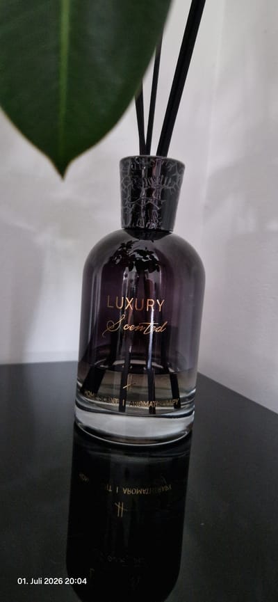 This is a close-up, rotated photograph of a luxury scented reed diffuser. The diffuser bottle is made of dark purple glass and has a textured, black lid. Inside the bottle, several black reeds are submerged in the liquid, ready to release fragrance. The bottle has gold lettering that reads "LUXURY Scented" on one side, and "HOME SCENE AROMATHERAPY" on another. The background is split between a plain white surface and a dark, possibly black, surface. A large, dark green leaf is partially visible in the lower left corner of the frame, suggesting it is placed near a plant. The lighting appears to be artificial. A timestamp in the bottom right corner reads "01. Juli 2026 20:04," indicating the photo was taken at 8:04 PM on July 1, 2026. There are no people visible in the image.