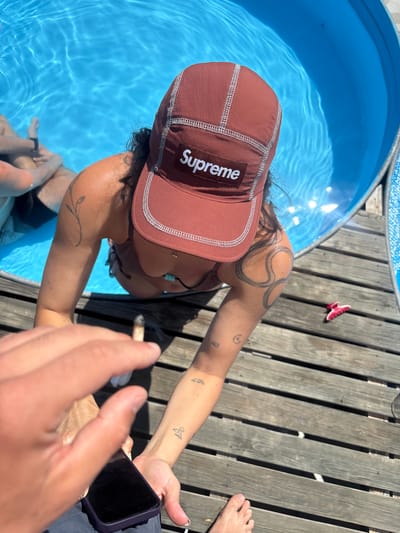 A person wearing a brown Supreme baseball cap and a brown bikini top is leaning into a circular blue pool filled with clear water. They have several tattoos on their arms, including a plant-like design on their left upper arm and a spiral design on their right upper arm. Another person, partially visible, is in the pool with them. The pool is surrounded by a wooden deck. A hand holding a smartphone is in the foreground, obscuring part of the scene. A pink hair clip is visible on the deck to the right of the pool. The overall setting appears to be an outdoor recreational area, possibly a backyard or a poolside area. The image does not provide any information to ascertain its location in San Lorenzo De Tormes, Spain.