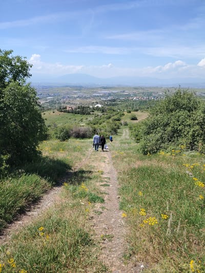 Two individuals are walking down a dirt path on a grassy hillside. The person in front is wearing a blue and white checkered shirt and dark pants, carrying a dark bag and a water bottle. The person behind them is wearing a dark outfit and carrying a white bag. They are walking away from the camera, towards a city visible in the distance. The hillside is covered in green grass, with scattered yellow wildflowers. Trees are visible on either side of the path, and in the background, a city sprawls across a valley with mountains beyond. The sky is blue with scattered white clouds. The scene appears to be in the vicinity of Jrvej, Armenia, given the topography and distant urban landscape often associated with its surrounding areas.