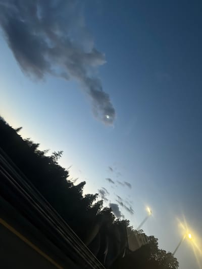 This is an angled, low-light, outdoor, wide shot of a sky over trees with streetlights. The sky is a gradient from light blue near the horizon to a deep blue at the top of the frame. There are scattered clouds, some dark and wispy, one larger cloud with a bright, circular object behind it that appears to be the sun or moon. The lower portion of the image is dominated by the dark silhouettes of trees and some structure, possibly a railing or barrier, in the foreground. Two bright yellow streetlights are visible in the upper right quadrant of the image, with beams of light extending diagonally upwards. The image appears to have been taken from a moving vehicle due to the motion blur of the trees. The location is Ås, Norway, suggesting this is likely a scene from the outskirts or a park area within the city as dusk or dawn approaches. There are no people or discernible actions taking place.