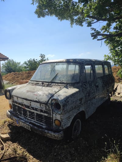 This media file shows an old, dilapidated white van parked outdoors in a rural setting. The van is the primary subject, appearing heavily weathered with peeling paint and rust. The front of the van is visible, revealing a damaged grille and a missing headlight. A license plate with the text "16 LU 940" is attached to the front bumper.

The van is situated on a dirt surface surrounded by dry grass and weeds. In the background, there is a pile of dirt or construction material and a glimpse of a building with a tiled roof. Trees with green foliage are present, casting shadows on the van and the surrounding ground. The sky is clear and blue, suggesting a sunny day. The overall scene evokes a sense of abandonment and disrepair. The lighting indicates it's daytime, likely mid-morning or afternoon due to the sun's angle and the shadows. The presence of the license plate with Turkish characters ("TR") and the location context of Osmangazi, Turkey, provide specific geographical information. No people are visible in the image, and no specific activity or event is taking place; the focus is on the static state of the abandoned vehicle.