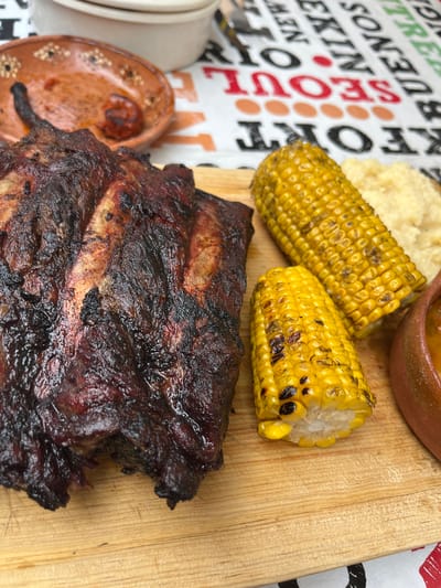 A close-up shot of a wooden board featuring food. The primary subject is a large portion of barbecued ribs, with a dark, crusty exterior. Beside the ribs are two pieces of grilled corn on the cob, slightly charred in places. A portion of what appears to be mashed potatoes or a similar side dish is also visible. Additionally, there are small ceramic bowls containing sauce. The background consists of a table covering with various city names, including "SEOUL" in red letters and "NEW YORK" and "PEKIN" in black. The location, San Francisco Totimehuacán, Mexico, cannot be determined from the image content.