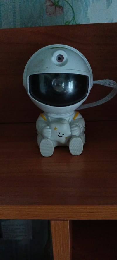 A close-up, eye-level, indoor shot captures a small, white astronaut figurine sitting on a wooden shelf. The figurine is designed to resemble a cartoon astronaut, complete with a helmet that has a large, reflective dark visor and a small red circular "eye" at the top. The astronaut is holding a white cloud-like object with a simple curved smile and two small yellow stars. The astronaut's suit is white with yellow accents on the shoulders. A grey, woven strap extends from the right side of the helmet, drooping slightly. The background consists of a wooden shelf surface and a blurred, patterned wallpaper with floral elements. The scene is dimly lit, suggesting it is within a home or storage area. The location is identified as Александровский Завод, Russia, though this specific image provides no direct visual cues to confirm this geographical context, other than what is provided in the prompt.