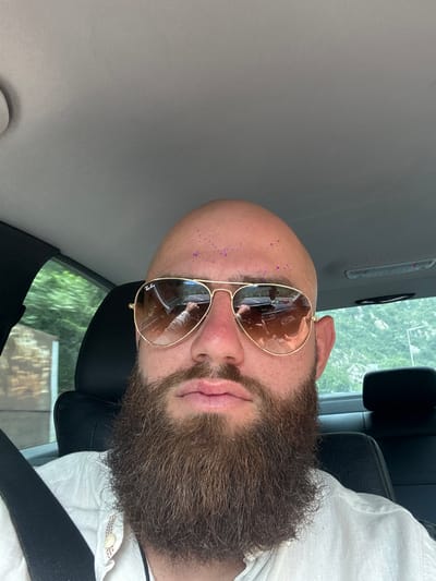The image shows a man with a bald head and a full beard wearing gold-rimmed sunglasses, sitting in a car. Purple glitter is visible on his forehead. He is wearing a white shirt and a black seatbelt. The interior of the car, including the grey roof and black seats, is visible. The reflection in his sunglasses shows a view of the car's interior and an arm holding a phone, suggesting the photo was taken from inside the vehicle. The location appears to be near greenery, but there is no indication that the location is in Krupnik, Bulgaria.