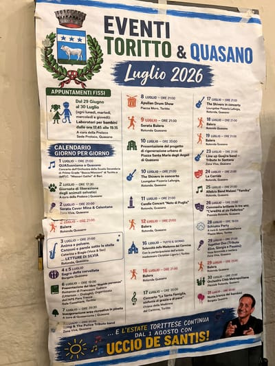 This is a photograph of a poster advertising events in Toritto and Quasano, Italy, in July 2026. The poster features a town crest with a bull and a laurel wreath at the top left. Below this, a section titled "APPUNTAMENTI FISS" (Fixed Appointments) lists ongoing activities. The main body of the poster is a "CALENDARIO GIORNO PER GIORNO" (Day-by-Day Calendar) detailing events from July 1st to July 31st. The events include concerts, performances, workshops, festivals, and religious ceremonies. Icons are used to represent different types of events, such as musical instruments, animals, and people dancing. The bottom of the poster has a banner with the text "...E L'ESTATE TORITTESE CONTINUA DAL 1 AGOSTO CON..." followed by the words "UCCIO DE SANTIS!" and an image of a smiling man pointing towards the viewer. The poster is taped to a wall, and the edges show signs of wear and tear.