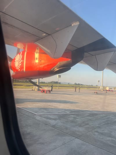 The image shows the wing and engine of a Lion Air airplane viewed from an interior window, likely taken at Baji Mangngai, Indonesia. The wing is light gray, with darker gray and black accents. The engine is bright orange, featuring a white lion logo. Outside the window, the airport tarmac is visible, constructed of light gray concrete. A grassy area separates the tarmac from what appears to be a distant mountain range or elevated terrain. Airport equipment and personnel are present on the tarmac. The sky is clear and light blue.