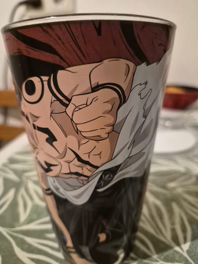 A close-up, slightly angled view shows a glass with an anime-style illustration of a muscular character with dark hair and a light-colored shirt. The character's fist is prominent in the foreground, with visible muscle definition and black markings on the arms and torso. The glass is placed on a table with a patterned green tablecloth. In the blurred background, a red bowl and white plates are visible. The scene is indoors, with the lighting suggesting it is daytime.