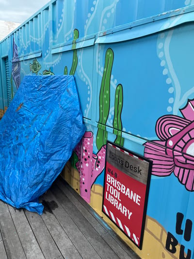 This outdoor image, likely taken in Hamilton, Australia, captures a brightly painted structure resembling a shipping container or building wall, adorned with an underwater-themed mural. The mural features stylized sea creatures and marine vegetation in vibrant blues, greens, pinks, and white, creating a lively visual.

A prominent red and white sign with a black frame is attached to the painted wall. The text on the sign reads "Loans Desk" on a grey section, followed by "W4-B", and then "BRISBANE TOOL LIBRARY" in large white capital letters against a red background with diagonal white stripes. Partially visible above this sign is more painted text on the structure, appearing to be "LI BU".

At the base of the painted structure, a large, crumpled blue tarp is spread out, covering an object or area beneath it. To the right, a section of wooden decking or flooring is visible, suggesting a public walkway or outdoor space. The lighting indicates it is daytime, with soft, diffused light. There are no people visible, and no active event is taking place, presenting a static view of a community-oriented facility.