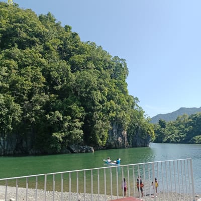 The image captures a scenic view of a lake surrounded by lush green hills under a clear blue sky. In the foreground, a white metal fence separates the viewer from the pebble-strewn shore and the calm, jade-colored water. Several people are visible near the water's edge and in the lake. Two individuals are in a kayak on the water, paddling. On the shore, at least three people, including two children, are standing near the water. The shoreline is lined with trees and rocky outcrops, and the backdrop is dominated by a densely forested hillside that slopes down to the lake. The overall scene suggests a tranquil outdoor recreational setting in a natural environment. The location is identified as the city of Lamseunia, Indonesia.