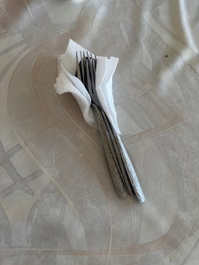 The image shows two forks wrapped in a white paper napkin, resting on a table with a patterned surface. The forks appear to be made of metal and have decorative handles. The napkin is folded and partially obscures the forks. The table surface has a light-colored pattern with curved lines and geometric shapes. The lighting suggests an indoor setting, possibly a restaurant or dining area. The location is identified as Kara-Suu, Kyrgyzstan.