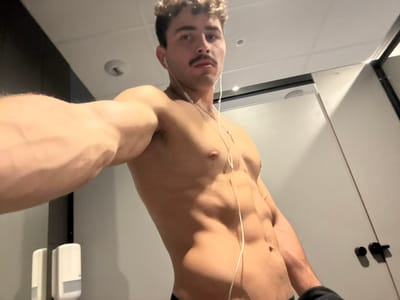 The scene depicts a shirtless male individual with a muscular physique, prominent biceps, and visible abdominal musculature, posing for a selfie in an indoor setting. He has curly brown hair and a distinct dark mustache, and is wearing white wired in-ear headphones. His right arm is extended into the foreground, flexing the biceps and showing prominent veins. The background features light-colored walls, with a white wall-mounted dispenser visible on the left. A white door with a black handle is partially visible on the right. The ceiling is white, likely tiled, and includes a circular recessed light fixture. This appears to be a locker room or public restroom facility, located in Chantepie, France.