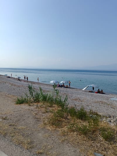 A wide, long shot captures a pebble beach along a calm, blue body of water under a clear, pale blue sky. The beach is lined with people relaxing under umbrellas and near the water. A dirt path with dry grass runs along the foreground. The distant horizon shows faint, hazy mountains.