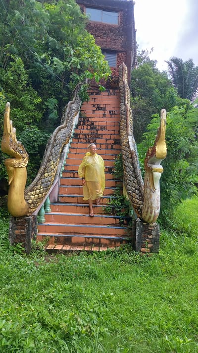 A person in a yellow raincoat is lying on an orange brick staircase flanked by two serpentine statues. The staircase leads up to a building with a stone facade. Lush green vegetation surrounds the structure, including grass on the right side of the frame and trees behind the building. The location is Thap Put, Thailand.