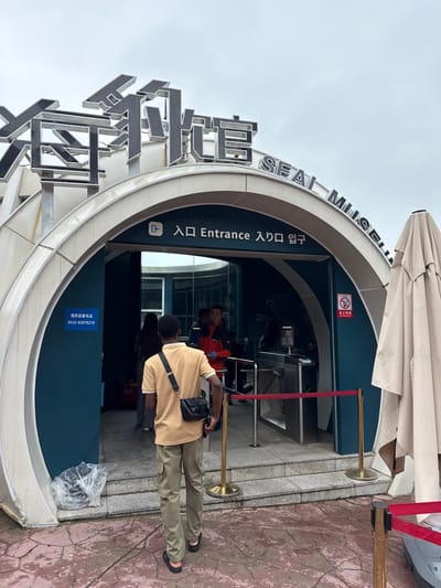 The image depicts the entrance to the Seal Museum (海豹馆 SEAL MUSEUM) located in Shinan District, China. The building features a modern, arched facade with metallic grey exterior panels and a deep teal interior section at the entrance. Above the entryway, large, dark grey Chinese characters for "海豹馆" are prominently displayed, followed by "SEAL MUSEUM" in English. Directly above the inner glass doors, a smaller sign in multiple languages reads "入口 Entrance 入口 입구".
In the foreground, a male individual with dark skin, wearing a light tan polo shirt, olive green cargo pants, and black sandals, walks away from the viewer towards the museum entrance. He carries a black shoulder bag. Inside the entrance, an automated stainless steel turnstile system is visible, preceded by red velvet ropes on brass stanchions, forming a queue line. Other individuals are visible beyond the turnstiles; one person wears a red and orange jacket. A blue sign on the left interior wall displays a Chinese service supervision phone number: "0532-82878218". On the right interior wall, a red and white "no smoking" sign is affixed. To the far right of the entrance, a large, beige, folded outdoor umbrella is partially visible. The ground is paved with reddish-brown, irregularly shaped flagstones under an overcast sky. A clear plastic bag containing trash is positioned near the base of the left arch.