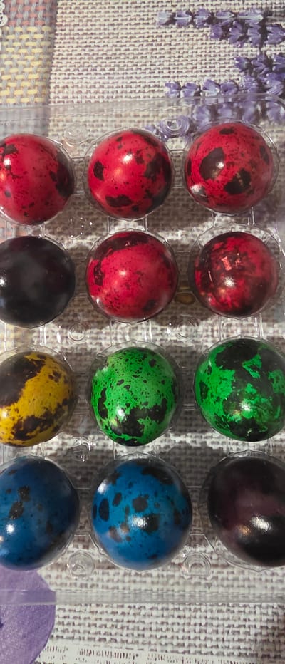 The image shows a carton of twelve quail eggs dyed in various colors, including red, green, purple, black, yellow, and blue. The eggs are speckled with darker spots. They are arranged in a clear plastic egg carton. The carton sits on a patterned surface that appears to be a tablecloth or mat with a floral design. The location is not discernable from the image alone.