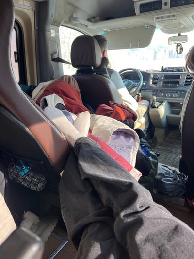 The image depicts the interior of a Volkswagen multi-passenger vehicle, likely a van, in Samolet, Russia, during daylight hours. The perspective is from a rear passenger seat, looking forward. In the immediate foreground, the legs of a person are visible, clad in dark grey pants and white crew socks, propped up over a pile of garments. This pile, consisting of various jackets in colors such as red, white, purple, and blue, occupies the space between the front seats. The driver, a male wearing a light-colored long-sleeved shirt, is visible from the rear, hands on the steering wheel bearing the Volkswagen emblem. A second passenger's head is partially visible on the far right. The vehicle's dashboard features a manual gear shifter and an infotainment unit. An air freshener hangs from the rearview mirror. On the left, a brown leather-like seat has a mesh pocket containing a blue object. A white furry slipper or boot is on the floor near the foreground passenger's feet. Through the front windshield, a brightly illuminated outdoor environment with sparse trees and a light-colored ground, possibly snow, is discernible.