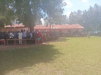 The image depicts an outdoor gathering in Idakho South ward, Kenya, under a clear sky. In the midground, a large group of individuals is assembled on a grassy field. Many are positioned near or on multiple wooden benches, which are arranged under the shade of a large, leafy tree on the left. Two single-story buildings with light-colored walls and reddish-brown corrugated iron roofs are visible in the background, behind the gathering. A dense line of tall, slender trees forms the distant background on the right. Individuals are dressed in varied civilian clothing. A smaller group on the far right of the assembly appears actively engaged, with some wearing more vibrant attire, suggesting a performance or ceremonial activity.