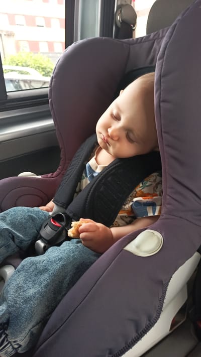 A baby sleeps soundly in a car seat, its head resting against the padded side. The baby is wearing a light-colored t-shirt with a playful pattern of animals and blue jeans. A small piece of food, possibly bread, is held in its left hand. The car seat is dark purple with black straps. The background shows a window with a view of a building with red and white striped facade and green foliage, suggesting a location in Altdorf bei Nürnberg, Germany. The lighting indicates daytime.