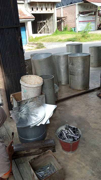 In Geulanggang Teungoh, Indonesia, a scene unfolds on a concrete surface likely outside a workshop or storage area. Several large, cylindrical metal containers are prominent in the mid-ground, appearing to be made of galvanized steel. To the left, a dark, corrugated metal gate stands partially open, revealing glimpses of a building under construction with a rusted corrugated metal roof and unfinished brickwork.

In the foreground, a person, partially visible from the shoulders down, is actively working. They are wearing a light-colored, patterned shirt and orange shorts. The person is holding a hammer in their right hand, poised to strike a piece of metal resting on a heavy, dark metal object, possibly a workbench or anvil. To the right of this workspace, a red bucket overflows with shiny, cylindrical metal pieces, likely components or finished products. Adjacent to the bucket, a large, square metal bin contains more of these metal pieces, neatly arranged.

Further left, a white plastic bucket, stained and weathered, sits inside a grey metal box. A bundle of thin, coiled metal wire is also present, resting on a stack of flat, circular metal sheets. The environment suggests an industrial or craft-oriented setting where metal fabrication or assembly is taking place. The background shows more of the unfinished building and surrounding vegetation.