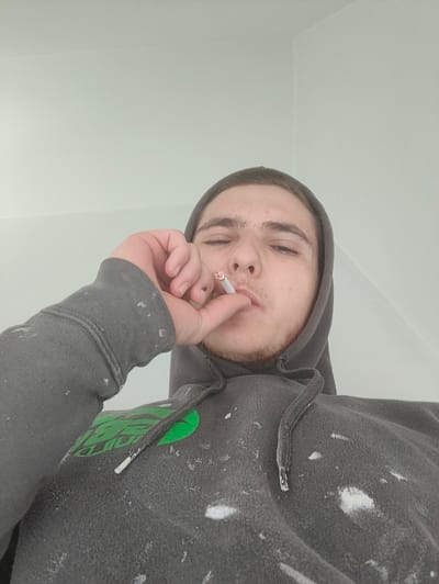 The image displays a young male subject captured from a low-angle perspective, engaged in smoking. He has short dark hair, light skin, and visible stubble on his chin and jawline. The subject is wearing a dark grey hooded sweatshirt adorned with a green logo on the left chest and numerous white paint splatters distributed across the fabric, including the sleeves and hood drawstrings. His left hand, showing a dark mark on a fingernail, holds a lit cigarette to his mouth; the cigarette's tip exhibits an orange-red glow. His eyes are partially closed. The background is an undecorated, light-colored interior wall, with a subtle corner visible in the upper right quadrant of the frame. This scene is located in the city of Deerlijk, Belgium.