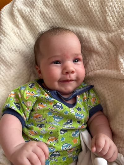 A close-up, eye-level shot shows a baby lying on a textured, cream-colored blanket. The baby is wearing a bright green onesie with a navy blue trim around the collar and sleeves. The onesie features a repetitive pattern of cartoonish cars in orange, blue, and black, along with stars and text that appears to say "Vrum Vrum" and "my happy car." The baby's head is slightly tilted to the right, with one eye looking towards the camera and the other slightly off to the side. The baby has fair skin, sparse brown hair, and plump cheeks. The lighting is natural, casting soft shadows. The background consists entirely of the soft, textured blanket. The location is not specified in the image or the provided hints.