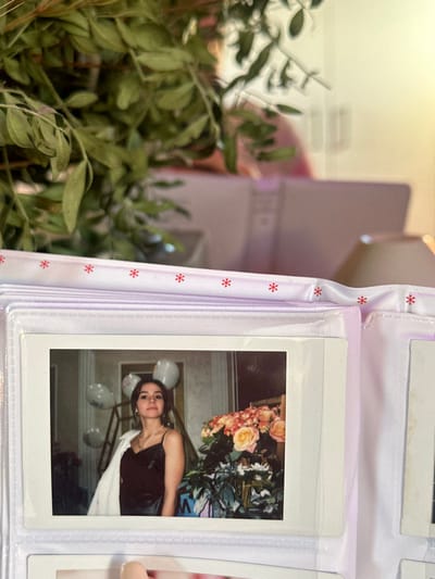 An instant print photograph, measuring approximately 7.5 cm by 10.5 cm, is displayed within a clear plastic sleeve of a light purple photo album. The album's upper edge features a white ribbon adorned with repeating red asterisk symbols. The photograph depicts a young female, approximately 20-30 years old, with dark hair and a neutral expression, looking directly at the viewer. She wears a black spaghetti-strap dress and has a white jacket draped over her left shoulder. A necklace with a pendant and earrings are visible. To the right of the subject is a large arrangement of light orange and pink roses with green foliage. In the background, to the left of the subject, several white balloons are visible. The setting within the photograph appears to be an indoor space with a dark wall. In the immediate foreground of the overall image, a dense cluster of green leaves and branches partially obstructs the upper portion of the photo album. The blurry background behind the album includes light-toned, indistinct objects, one of which appears to be a pink laptop or folder, and another, a light-colored lamp. The scene is located in 3-Y Uchastok, Russia.