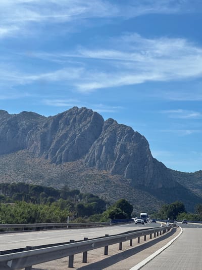 The image shows a view from a highway in Ondara, Spain, featuring a mountainous landscape under a blue sky with wispy clouds. The road, seen on the right, has a metal guardrail and light-colored asphalt with white lane markings. Traffic is backed up on the highway. A large mountain is centered, covered in rocky terrain and sparse vegetation.
