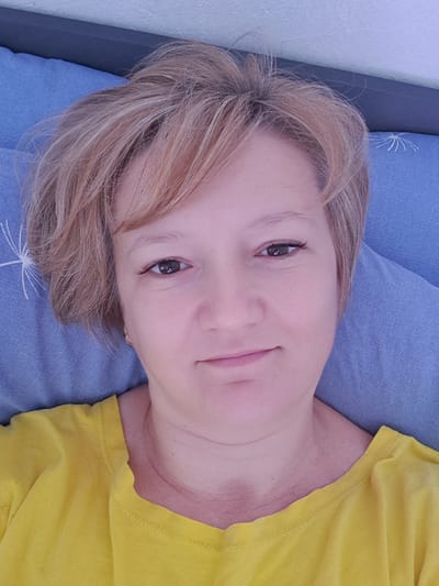 The image is a close-up, overhead selfie of a woman with short, blonde hair, lying down. She is wearing a yellow t-shirt. Her face is the primary focus, with her eyes open and a slight smile. The background consists of a blue pillow with a white pattern. The lighting appears natural, suggesting an indoor setting. The image was taken in Khadyzhensk, Russia.
