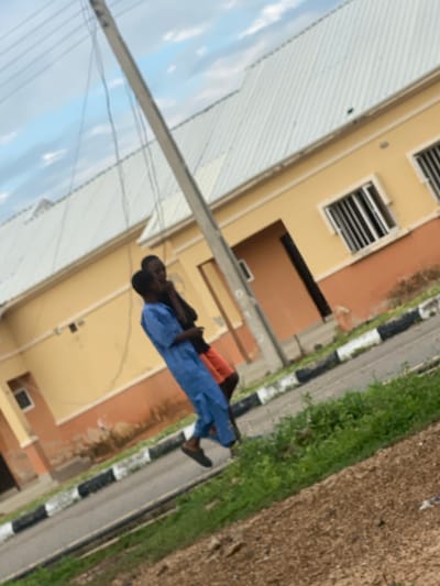 In Dumnga, Nigeria, two individuals are captured in a photograph taken outdoors. A man in a blue garment stands next to another person, partially obscured. They are positioned near a yellow and brown building with a white corrugated roof. A utility pole with wires runs vertically through the frame, angled slightly. The ground in front of the building is paved with a road marked with black and white curbs, bordered by green vegetation and brown soil. The sky above is partly cloudy.