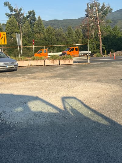 The image shows a street scene in Simitli, Bulgaria. In the foreground, a grey car is parked on a gravel surface. Behind the car, there is a raised planter with some greenery. Further in the background, two orange trucks are parked. One truck has a flatbed, and the other appears to be a utility van. A tall, dead tree stands to the right of the trucks. The background consists of lush green trees and rolling hills under a clear blue sky. There are traffic signs and a red circular sign with a diagonal bar visible near the road. A dog is seen walking on the side of the road. The scene appears to be a roadside area with some construction or maintenance vehicles present.