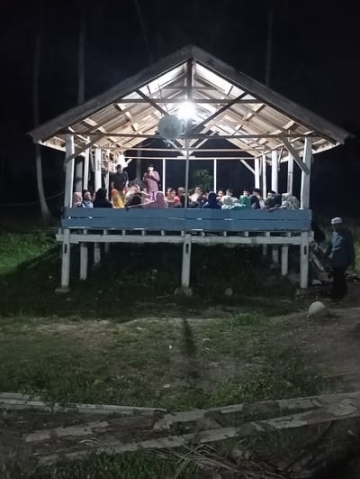 In Aluekeujruen, Indonesia, at night, a group of people are gathered under a wooden, open-sided pavilion. The pavilion is illuminated by a bright light hanging from its ceiling, casting shadows on the wooden beams and the people below. The structure is elevated on white pillars and has a blue railing along its front.

Inside the pavilion, approximately 15 to 20 individuals are seated, facing towards the center where two men are standing. One standing man is wearing a red shirt and holding a microphone, appearing to be speaking. The other man, in a dark shirt, stands beside him. The seated individuals appear to be a mix of genders and ages, some wearing headscarves.

Outside the pavilion, to the right, a lone figure in a white hat and dark clothing stands facing away from the structure. The ground surrounding the pavilion is covered in grass and dirt, with some fallen palm fronds visible in the foreground. Tall, dark trees are silhouetted against the night sky behind the pavilion. The overall scene suggests an evening gathering or event taking place outdoors.