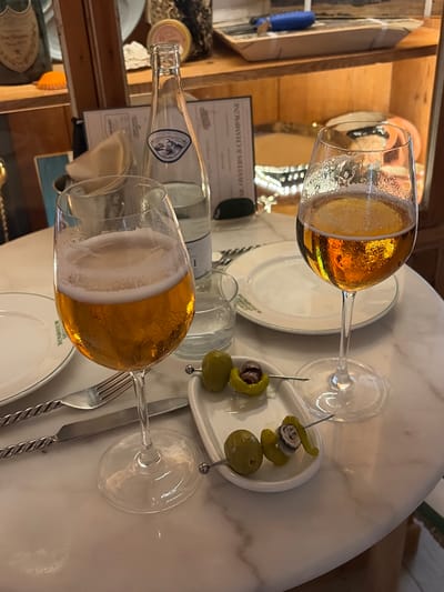 The image depicts a table at a restaurant, likely in Maó, Spain, with two glasses of amber-colored liquid, possibly beer or wine, and a bottle of water. The glasses are on a white marble-topped table. In the foreground, there are two skewers with olives and what appears to be anchovies or small seafood pieces. The table is set with silverware, including a fork and a knife with twisted handles. In the background, shelves are visible, holding various items including what seems to be a bottle of champagne or sparkling wine with a prominent label, and other decorative objects. A menu card is also visible, and part of its text reads "OSTRS & CHAMPAGNE". The overall scene suggests a casual dining or aperitif setting.