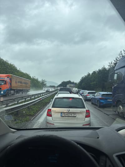 The image shows a traffic jam on a highway in Gorna Vasilitsa, Bulgaria on a rainy day. Multiple cars and trucks are seen stopped in both lanes. The car directly in front of the camera is a white Skoda Octavia with Bulgarian license plate PB 4074 TP. The sky is overcast. A mountain is visible in the distance. A truck with the words "HEPPER & OCHSENDORFER" is visible in the opposite lane.