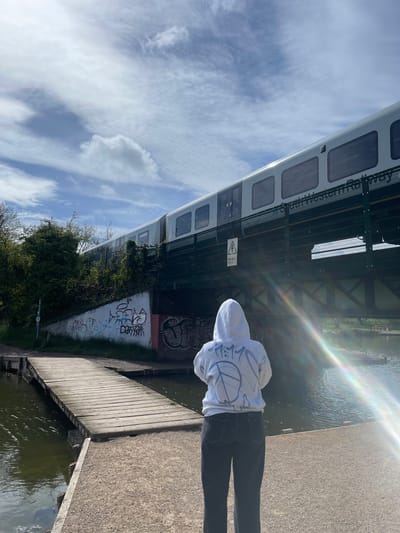 The image captures an outdoor scene featuring a person standing on a paved pathway next to a body of water, with a railway bridge and train overhead. The perspective is from behind the individual, who is wearing a white hoodie with a prominent peace sign and other dark graffiti-style designs on the back, paired with dark trousers.

To the person's right is a calm body of water, likely a river or canal, reflecting the sunlight. A wooden boardwalk or jetty extends along the water's edge. The pathway they are standing on appears to be made of gravel or small stones.

Above and slightly to the left, a railway bridge spans across the scene. A white train carriage, belonging to "South Western Railway," is visible on the bridge. The bridge structure is made of metal girders and includes fencing. Underneath the bridge, concrete walls are covered with various, largely illegible graffiti tags. Dense green foliage and bushes grow along the bank beneath the bridge.

A yellow warning sign featuring an electrical hazard symbol (lightning bolt in a triangle) and the word "Danger" is affixed to the bridge structure.

The sky is bright and blue with scattered white clouds, indicating a clear, sunny day. A strong sun flare or lens flare with rainbow hues emanates from the upper right portion of the image, suggesting the sun is shining brightly from that direction. The overall impression is a vibrant, sunny day in an urban setting, possibly along a waterfront path in Portsmouth, United Kingdom.