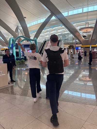 The image displays a brightly lit, expansive interior of a modern airport terminal in Al Matar, United Arab Emirates. The floor consists of large, reflective light-colored tiles, mirroring the high, geometrically paneled ceiling and the ambient light. Two males are centrally positioned in the mid-ground, walking away from the camera. The male on the left, with light-colored hair, wears a white short-sleeved t-shirt bearing 'THE NORTH FACE' logo on the back, dark trousers, and white sneakers; his left arm is raised and extended with the index finger pointing upwards and to the left. The male on the right, wearing a brown baseball cap, white short-sleeved t-shirt, dark trousers, and dark sneakers, carries a dark roll-top backpack and pulls a small, dark wheeled suitcase with his right hand. To the left of the central figures, a male in dark attire stands facing right, near a light-colored counter. In the background, an escalator ascends on the left, adjacent to teal-colored decorative architectural structures. A large, ornate circular chandelier hangs from the ceiling in the mid-right. Further in the background, digital display screens and a white directional sign indicating '← B C E F' are visible, alongside numerous blurred individuals navigating the terminal space.