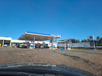 The image shows an Astral petrol station, taken from inside a car. The location is on the outskirts of Nduma, Kenya at latitude -1.172 and longitude 36.641. The station is surrounded by a white fence and has a blue and white awning. There are four gas pumps with vehicles parked at them. There is also a small building on the left side of the image, and a sign on the right side with the Astral logo. The sky is blue and clear. There are several power lines strung across the sky, and several green trees and bushes in the background.
