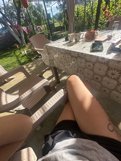 The image depicts a sunlit outdoor scene on a patio in Rogozen, Bulgaria. A table covered with a floral-patterned tablecloth is set with a coffee mug that reads "Coffee It's Coffee Time", a packet of cookies, a phone, and a glass. Two beige plastic lounge chairs are visible in the foreground, one of which is partially occupied by a person's legs. The legs are tanned and adorned with a tattoo on the right thigh. Behind the table, a fence encloses a garden with blooming red roses and lush green foliage. In the background, a red car is parked under trees. The overall impression is a relaxed, domestic setting enjoyed on a warm day.