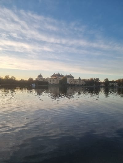 The image displays Drottningholm Palace, located on Lovön island in Ekerö kommun, Sweden, viewed across a body of water. The prominent subject is the light-colored Baroque palace, featuring multiple symmetrical wings, numerous windows, and several domed roofs, positioned on the far shore. A line of trees with mixed green and light autumn foliage partially obscures the palace's base. A small white boat is present on the water to the left of the palace structure. The water surface is characterized by gentle ripples, providing a distorted reflection of the sky and the palace. The upper portion of the frame shows a blue sky with scattered, wispy white clouds. A warm, golden light is visible on the horizon towards the left, indicating either sunrise or sunset. No individuals, discernible actions, or interactions are present in the scene.