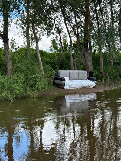 The image shows a discarded grey sofa partly covered with a white sheet, positioned on the riverbank in Izh-Boyki, Russia. Next to it is an old chair. The surrounding environment includes trees, bushes, and the water of the river. The reflection of the sky is visible in the water.