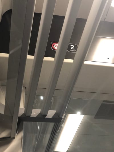 The image captures an interior view, likely within a modern public transportation setting such as a train, station, or ferry, characterized by clean lines and metallic finishes. The foreground is dominated by several parallel, horizontal bars, appearing to be made of a light gray or metallic material. These bars are evenly spaced, suggesting they are part of a railing, barrier, or window blind system.
Through the gaps in these bars, elements of the background are visible. On the left side, a dark, possibly black, rectangular sign or panel is partially seen. On this panel, a white circular emblem contains the number "2" and the word "classe" written vertically beside it, indicating "second class." Below this, a portion of another circular sign is visible, which appears to be a red prohibitory symbol (a circle with a diagonal line through it) with a white icon inside it, possibly depicting a person or a specific object, although it is too obscured to identify clearly. This sign might indicate a restriction like "no standing" or "no disabled access" depending on the full icon. The immediate background to the left of the dark panel is a light-colored wall or partition.
Towards the right side of the image, through and beyond the bars, more light gray or metallic panels are visible, some exhibiting subtle reflections. A prominent horizontal light fixture, emitting a bright white light, runs along the upper right portion of the scene. Reflections of this light source and other ambient lights are scattered across the metallic surfaces, adding to the bright and modern feel of the space. In the upper right corner, a section with a fine mesh or grid pattern is discernible, possibly a ventilation grate or part of a ceiling panel.
The horizontal bars in the foreground are supported on the right by a vertical structure, also in a darker gray or metallic tone, to which they are attached via white or light-colored brackets. The lighting is entirely artificial, creating a somewhat stark but well-lit environment. The overall impression is one of functionality and contemporary design, common in modern public transport or facilities.