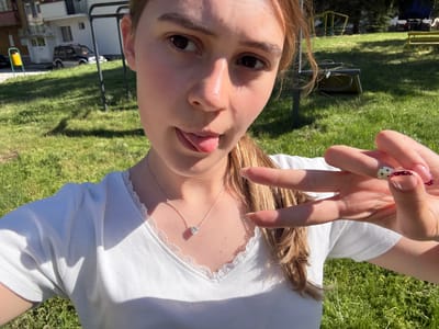 A young woman is lying on green grass in Panagyurski kolonii, Bulgaria. She is wearing a white top with lace trim around the neckline and a silver necklace with a pendant. She is holding up her left hand with her fingers in a peace sign, showing two manicured nails with decorative patterns. Her tongue is sticking out slightly. In the background, there are trees, playground equipment, a house, and cars.