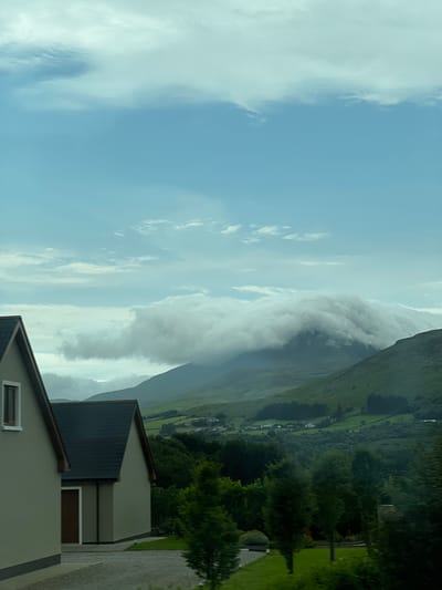The image depicts a cloudy sky over a lush green mountainous landscape. In the foreground, two modern houses with grey facades and dark roofs are visible on the left side. The houses appear to be residential properties. The background features rolling hills covered in vibrant green vegetation, dotted with scattered white houses. A large mountain in the mid-ground is partially obscured by dramatic, low-hanging clouds. The overall scene suggests a rural or semi-rural setting. The location is identified as Creeslough, Ireland. There are no people visible in the image.