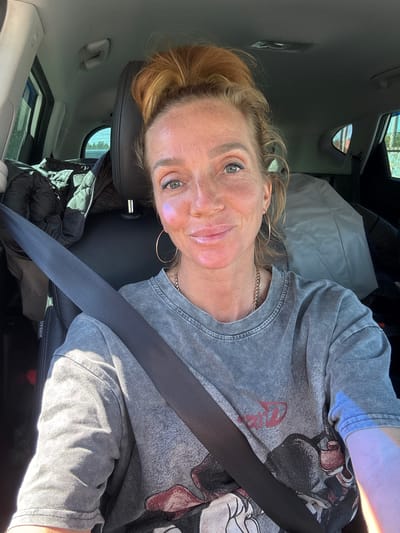A female individual is situated in the front passenger seat of a vehicle in Beloostrov, Russia. She is light-skinned, with numerous freckles visible across her nose and cheeks. Her hair is reddish-blonde, styled into a high bun atop her head. Her eyes are light-colored, appearing blue or grey, and she has dark eyeliner applied. A subtle, iridescent purple-blue highlight is visible on her left cheekbone. She maintains a closed-mouth smile and is looking directly forward towards the camera.

She is wearing a grey, short-sleeved t-shirt with a washed or distressed texture, featuring a partially visible graphic design that includes red and dark elements. A gold-toned chain necklace is around her neck, and she wears gold-toned hoop earrings. A black seatbelt is correctly fastened across her chest and lap.

The vehicle's interior is dark, with black leather upholstery on the headrests and black fabric on the seats. A black padded jacket is folded and placed on the rear seat behind her. Further back on the rear seat, a large, light grey object, possibly a blanket or bag, is visible. The word "AIRBAG" is visible on the pillar near her left shoulder. The scene is illuminated by strong daylight, indicating sunny conditions. Through the vehicle windows, blurred outdoor elements such as foliage, trees, and distant structures are discernible.