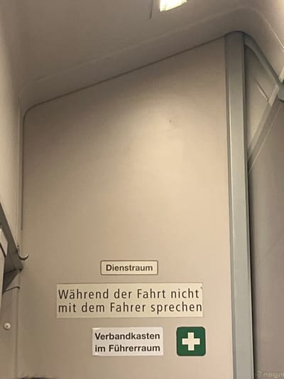 The image displays a section of a train's interior. A sign indicates "Dienstraum" (service room). Adjacent to this, another sign reads "Während der Fahrt nicht mit dem Fahrer sprechen," prohibiting interaction with the driver during the journey. A green cross denotes a first aid kit ("Verbandkasten im Führerraum"), located in the driver's cabin. The background consists of a smooth, neutral-colored surface, possibly a door or wall, with ceiling and framing elements visible.