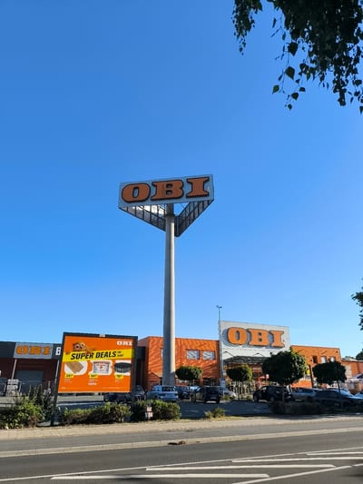 The image shows an OBI DIY store in Bremerhaven, Germany. A tall pylon sign with the OBI logo is prominently displayed against a clear blue sky. The store itself has an orange and grey facade with the OBI logo above the entrance. In front of the store, a billboard advertises "Super Deals im Juni!". Several cars are parked in the lot and on the street in front of the store. Trees and bushes are planted in front of the building and along the sidewalk. The overall scene depicts a retail environment on a sunny day.