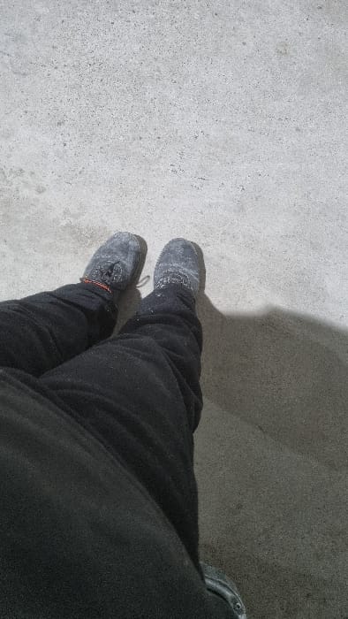 The image shows a pair of feet wearing dark gray or black moccasin-style shoes. The shoes appear worn and dusty. The person is wearing black pants. They are standing on a concrete surface that is lighter in color, likely a sidewalk or floor. A shadow is visible near the upper right portion of the image.