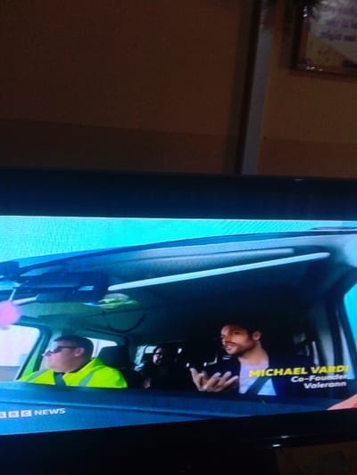This photograph, captured in Mista Ali, Nigeria, depicts a television screen broadcasting a news segment. The screen displays the interior of a vehicle, likely a van or SUV. A male driver, wearing sunglasses and a high-visibility yellow/green vest, is visible on the left, looking forward. In the rear passenger seat on the right, a man identified by an on-screen graphic as "MICHAEL VARD, Co-Founder Valerann," is shown speaking or gesturing with both hands open, palms up. He is wearing a dark shirt and a seatbelt. A third individual is partially visible in the rear on the left. The "BBC NEWS" logo is present on the lower-left portion of the screen's content. The environment outside the television screen includes a beige wall with a horizontal molding and a framed item in the upper right background.