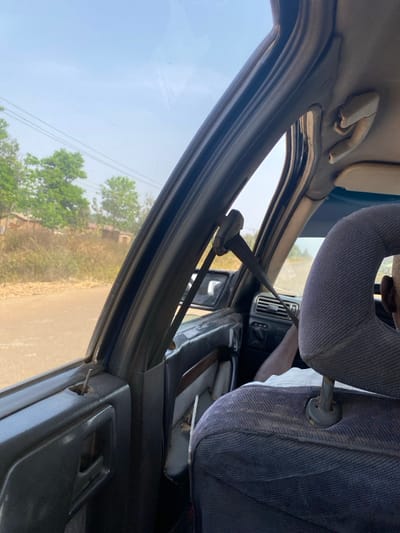 The scene depicts the interior of a vehicle, viewed from a rear passenger seat looking forward and slightly to the right, in Unguwar Gaya, Nigeria. The front passenger, a person with dark skin and short dark hair, is seated with their back to the viewer, wearing a light-colored shirt. Their right arm is visible. The seatbelt is extended from its B-pillar anchor point, but its use by the passenger is not discernible.
The vehicle's interior shows signs of wear: the dark blue/grey fabric of the front passenger headrest and the visible portion of the rear seat are soiled. The dark grey plastic door panel on the passenger side has a brown trim insert, and a metal rod protrudes from a hole where the door lock pin would typically be. The sun visor is folded up against the light-colored, worn headliner. A dashboard air vent is visible on the passenger side.
Outside the vehicle, a reddish-brown unpaved road is visible. Beyond the road, the environment consists of dry, sparse vegetation and several green trees with full foliage. In the mid-distance, indistinct brick-colored structures are partially visible. Overhead utility lines span across a clear, light blue sky. The overall impression is that of a vehicle traveling or parked on a rural or semi-urban road during daytime.