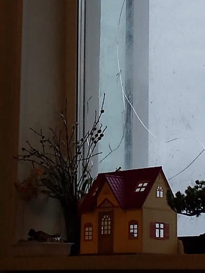 The image depicts a miniature, yellow house with a red roof, featuring several windows, some of which are illuminated. The house is positioned on a surface that appears to be a shelf or windowsill, with a bouquet of dried branches and pussy willows in a vase to its left. Behind the house and bouquet, a window is visible, with visible cracks on its surface. To the right of the house, a dark green, miniature tree is present. The lighting suggests it is either daytime with overcast skies or nighttime. The scene is indoors, with the window likely looking out. There is no specific indication of the city of Magadan, Russia, in the image itself; the location information is provided contextually. The overall impression is a still life arrangement, possibly decorative.